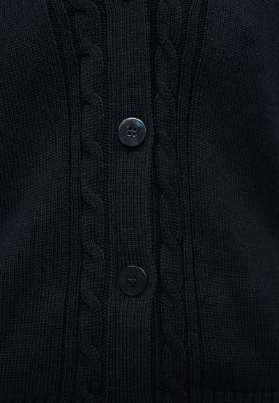 Σκούρα ναυτικά μπλε πλεκτή ζακέτα με δύο ορατά μαύρα κουμπιά με τέσσερις τρύπες και σχέδιο πλέξης με καλώδιο που τρέχει κάθετα δίπλα στα κουμπιά.