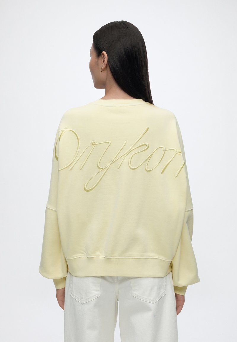 Femme aux longs cheveux foncés portant un sweat-shirt ample jaune pâle avec une inscription en relief dans le dos et un pantalon blanc, de dos.