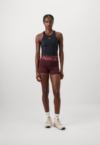 Čierny športový top s ružovými akcentmi, kombinovaný s bordovými šortkami s veľkým logom "NIKE". Model stojí na neutrálnej pozadí.