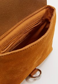 Portafoglio in suede color nocciola caldo con una texture morbida, dotato di diversi scomparti per carte e un anello metallico circolare. Interno foderato in tessuto chiaro.