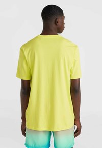 Camiseta de algodón amarilla con un corte relajado y mangas cortas, mostrada desde la parte de atrás. Combinada con pantalones cortos con degradado azul-verde.