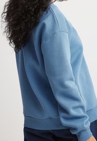 Persona con cabello rizado que lleva un sudadera ligera de color azul claro y jeans oscuros contra un fondo liso.
