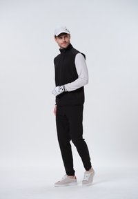 Zwarte mouwloze vest over een langemouwen wit shirt, gecombineerd met zwarte broek en grijze sneakers. Witte pet en golfhandschoen verfraaien de outfit.