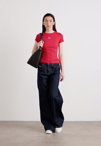 Une femme en chemise rouge et en jean large foncé s'approche de l'objectif, portant un sac à main noir sur un fond clair.