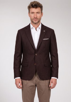 Man standing wearing a dark brown blazer, white dress shirt, beige trousers, and a patterned pocket square on a plain light background.