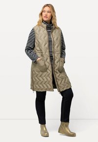 Wypchana, jasnooliwkowa kamizelka bez rękawów z kieszeniami bocznymi i zapięciem na zamek, noszona na pasku w prążki, w połączeniu z ciemnymi dżinsami i złotymi botkami.