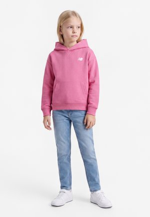 Pige iført en pink hættetrøje, lyseblå jeans og hvide sneakers, der står mod en ensfarvet baggrund og kigger til siden.