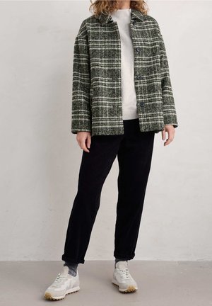 Person trägt eine grün-weiß karierte Jacke, einen leichten Pullover, schwarze Hose, graue Socken und weiße Turnschuhe und steht vor einem einfarbigen Hintergrund.