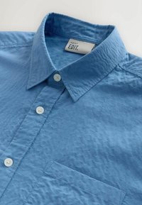 Chemise bleu texturée à boutons avec col, poche poitrine, et boutons blancs, présentant l'étiquette de la marque "NEXT EDIT." à l'intérieur du col.