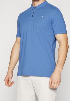 Mężczyzna w niebieskiej koszulce polo z krótkim rękawem z logo "CK" na piersi i jasnych spodniach, stojący na jednolitym tle.