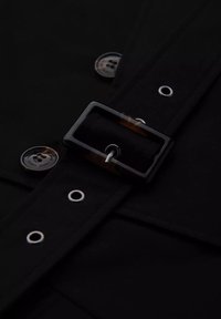 Czarna tkanina z detalami w postaci prostokątnej klamry, srebrnego guzika i metalowych oczek, podkreślająca elegancki, stonowany design.