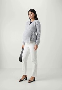 Cardigan de maternité en tricot gris avec fermeture à nouer, porté sur un haut sombre, associé à un pantalon blanc et des sandales à talons noirs.