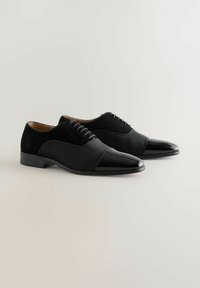 Paar schwarzer formeller Herrenschuhe mit glänzenden Zehenkappen, strukturierten Seitenpanelen und Schnürdesign auf hellem Hintergrund.