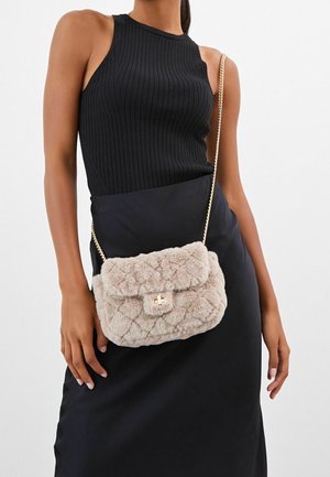 Nainen pukeutunut mustaan hihattomaan toppiin ja hameeseen, kantaa beigeä tikattua tekoturkisristikkolaukkua, jossa on kullanvärinen ketjusangallinen ja -lukollinen olkahihna.