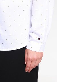 Camisa blanca de manga larga con un patrón texturizado de pequeños diamantes dorados, con un logo de Tommy Hilfiger en rojo y azul en el puño.