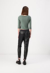 Femme aux cheveux bruns portant un top court vert, un pantalon en cuir noir fuselé et des chaussures à plateforme noires, se tient face à un mur blanc.