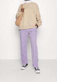 Felpa oversize beige con una patch logo, abbinata a pantaloni dritti lilla e sneakers bianco e nero, tenuta con una borsa beige.