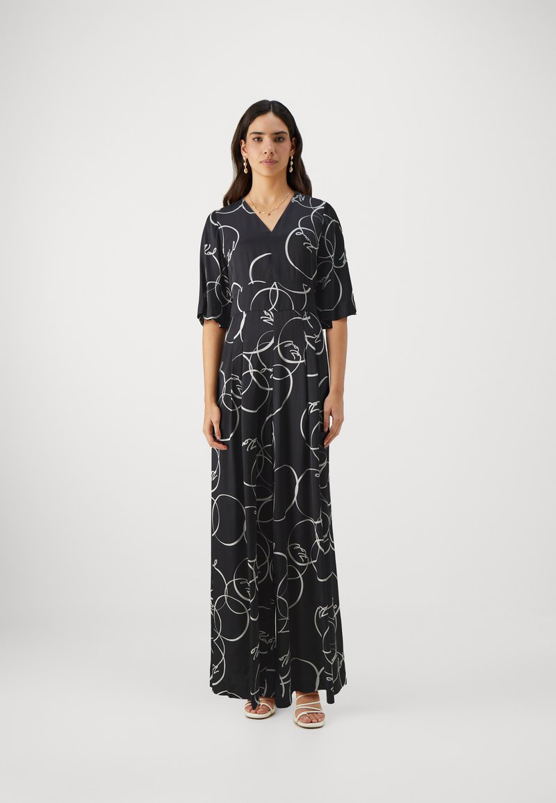 Black maxi dress with white abstract line patterns, V-neck, short sleeves, and a flowing silhouette. Paired with strappy white sandals.