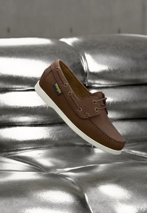 Καφέ δερμάτινο casual μοκασίνι με λευκή σόλα και κορδόνια, να αιωρείται πάνω από μια λαμπερή ασημένια επιφάνεια με καπιτονέ σχεδιασμό.