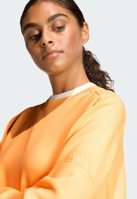 Pomarańczowa bluza z okrągłym dekoltem, wykonana z teksturowanej tkaniny i wyróżniająca się charakterystycznym elementem na rękawie. Gładkie wykończenie, luźny styl.