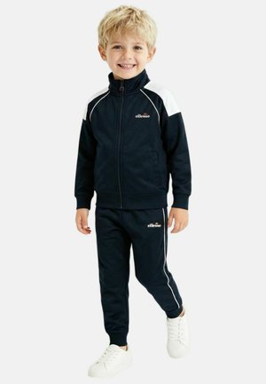 Jeune garçon souriant, marchant, portant un survêtement zippé noir et blanc avec un logo et des baskets blanches sur un fond uni.