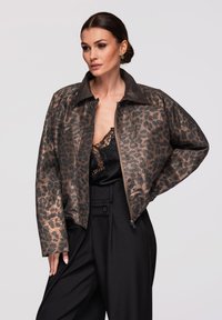 Femme portant une veste en cuir imprimé léopard marron, un caraco en dentelle noire et un pantalon noir taille haute, debout avec une main sur la hanche.
