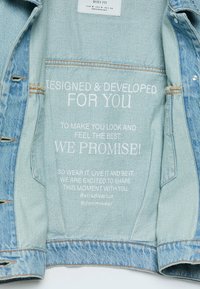 Giacca in denim azzurro chiaro aperta al rovescio che mostra un testo bianco: "Progettata e Sviluppata per Te... lo promettiamo! #stradivarius #denimwear."