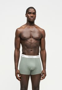 Hombre negro musculoso de pie mirando al frente, vestido con bóxers Calvin Klein gris claro con cinturilla blanca, fondo neutro.