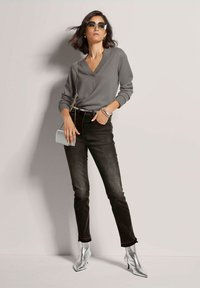 Grauer V-Ausschnitt-Pullover, schwarze Jeans mit schmalem Schnitt und glänzenden silbernen Stiefeletten. Die Accessoires umfassen eine kleine Handtasche und Sonnenbrillen.