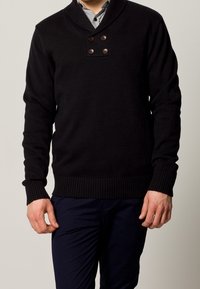 Svart stickad tröja med sjalkrage, prydd med två knappdetaljer vid halsen och ribbade ärmslut och nederkant.