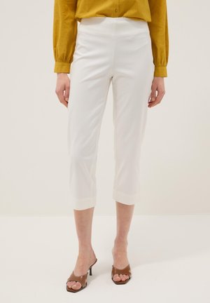 Vrouw draagt cropped witte broek gecombineerd met een mosterdgele blouse met lange mouwen en bruine open-toe sandalen met hak.
