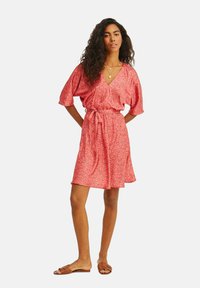 Šaty s květinovým vzorem v růžovém materiálu, krátké rukávy, v-výstřih a detail s uvázaným pasem. Obuví jsou hnědé sandály.