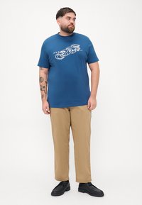 Modrá polo tričko s krátkým rukávem a bílým grafickým designem, spárované s khaki kalhotami a černými botami, na stojícím mužském modelu.