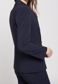 Blazer blu navy con design su misura, maniche lunghe e chiusura a un bottone. Tessuto liscio e silhouette strutturata.