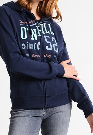 Kvinde iført navyblå O'Neill hættetrøje med lynlås og hætte samt lyseblå og pink bogstaver, kombineret med grå jeans, poserer med den ene arm på tværs af kroppen.