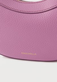 Piccola borsa a mano in pelle viola con finitura testurizzata, parte superiore curva e logo dorato discreto. Design compatto con accessori minimali.