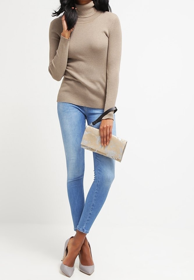 Femme portant un pull à col roulé beige, un jean skinny bleu clair, des talons hauts gris, tenant une petite pochette beige avec une sangle noire.
