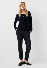 Navy cardigan dengan kancing, dipadukan dengan celana hitam dan loafers hitam beraksen emas, berlatar belakang putih polos.