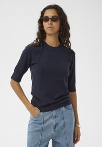 Marineblauer, gerippter Strickpullover mit kurzen Ärmeln, Rundhalsausschnitt und figurbetonter Silhouette. Kombiniert mit hellblauen, hoch taillierten Jeans.
