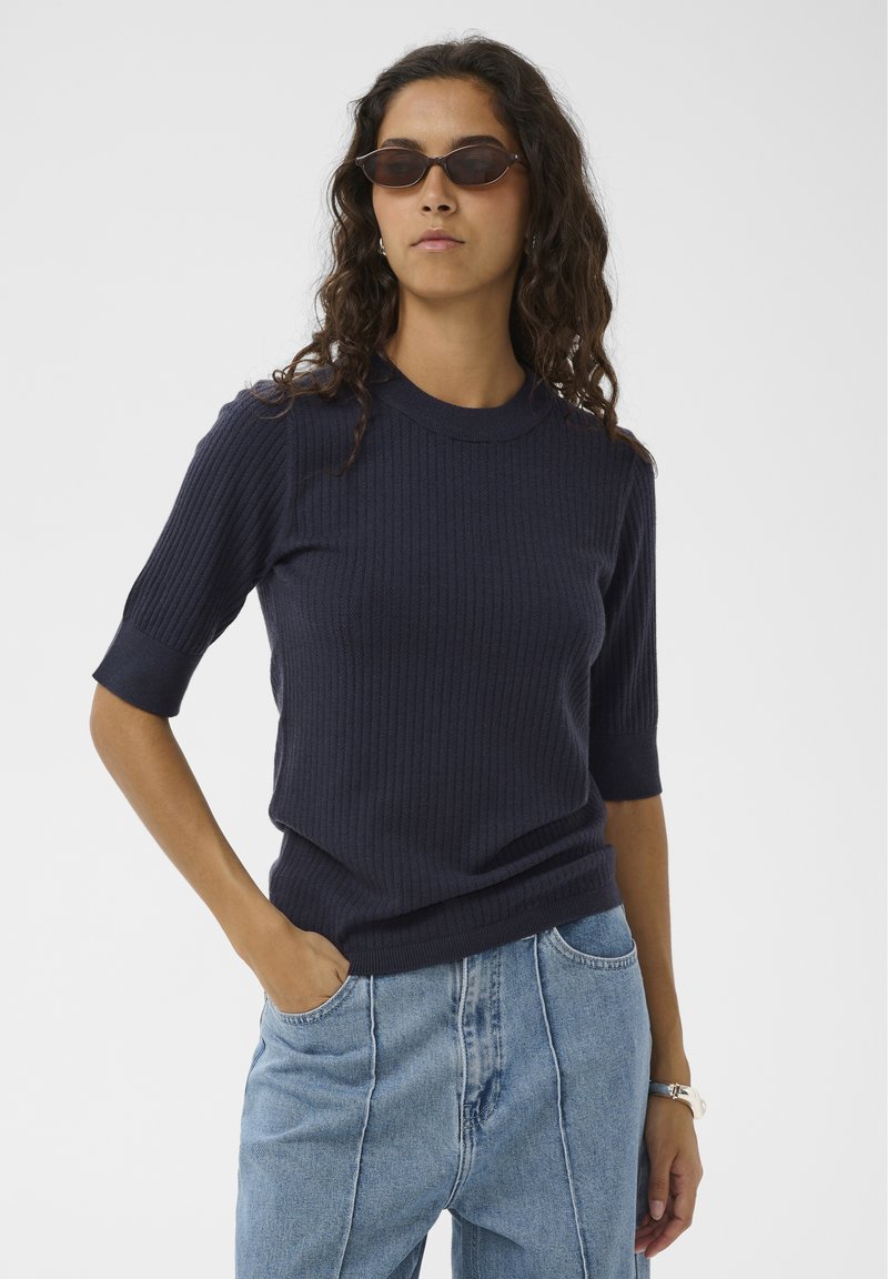 Marineblauer, gerippter Strickpullover mit kurzen Ärmeln, Rundhalsausschnitt und figurbetonter Silhouette. Kombiniert mit hellblauen, hoch taillierten Jeans.