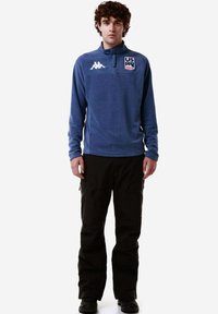 Pullover in pile blu con zip, logo degli Stati Uniti sul petto ed emblema Kappa. Abbinato a pantaloni da sci neri con tasche con zip.