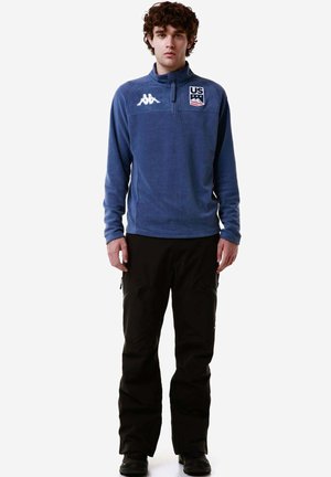 Pullover in pile blu con zip, logo degli Stati Uniti sul petto ed emblema Kappa. Abbinato a pantaloni da sci neri con tasche con zip.