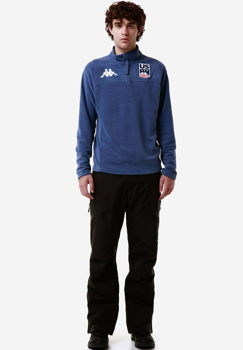 Pullover in pile blu con zip, logo degli Stati Uniti sul petto ed emblema Kappa. Abbinato a pantaloni da sci neri con tasche con zip.