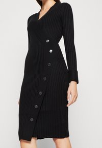 Donna che indossa un vestito avvolgente a coste nero con maniche lunghe e grandi bottoni argentati lungo il pannello frontale, in piedi di fronte a uno sfondo neutro.