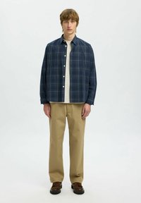 Camicia a quadri navy con bottoni bianchi, indossata sopra una T-shirt chiara, abbinata a pantaloni beige e scarpe marroni.