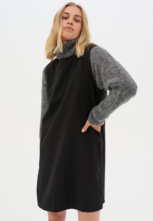 Schwarzes Kleid kombiniert mit grauen Strickärmeln. Rollkragen-Design, glattes Material und seitliche Taschen. Schlichte Silhouette mit strukturellem Kontrast.