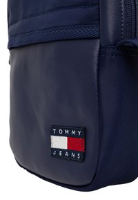 Mochila azul marino con una textura suave, que presenta un parche de logo rectangular con "TOMMY JEANS" y una combinación de colores rojo, blanco y azul.