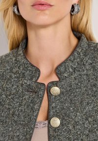 Vrouw in een grijze textuurjacket met decoratieve gouden knopen en zilveren oorringen, met blond haar en een top met kantafwerking zichtbaar.