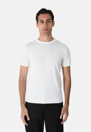Uomo con capelli corti e scuri che indossa un maglione bianco a maniche corte e pantaloni neri, in piedi davanti a uno sfondo bianco.