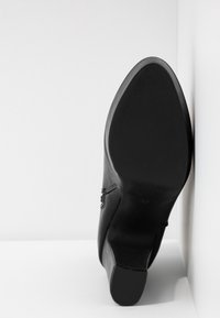 Černá kožená kotníková bota položená podrážkou nahoru v bílé rohu, ukazující hladkou, mírně texturovanou podrážku a nízký blokový podpatek.